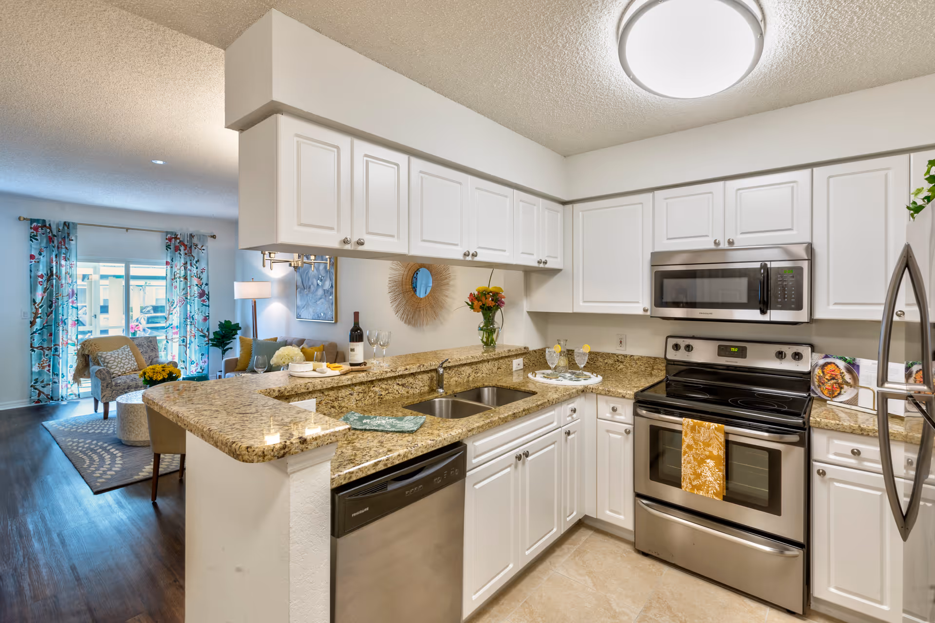Modern kitchen with white cabinets, granite countertops, stainless steel appliances including a stove, microwave, dishwasher, and refrigerator. The kitchen opens to a living area with a chair, a sofa, a floor lamp, and floral curtains on a sliding glass door.