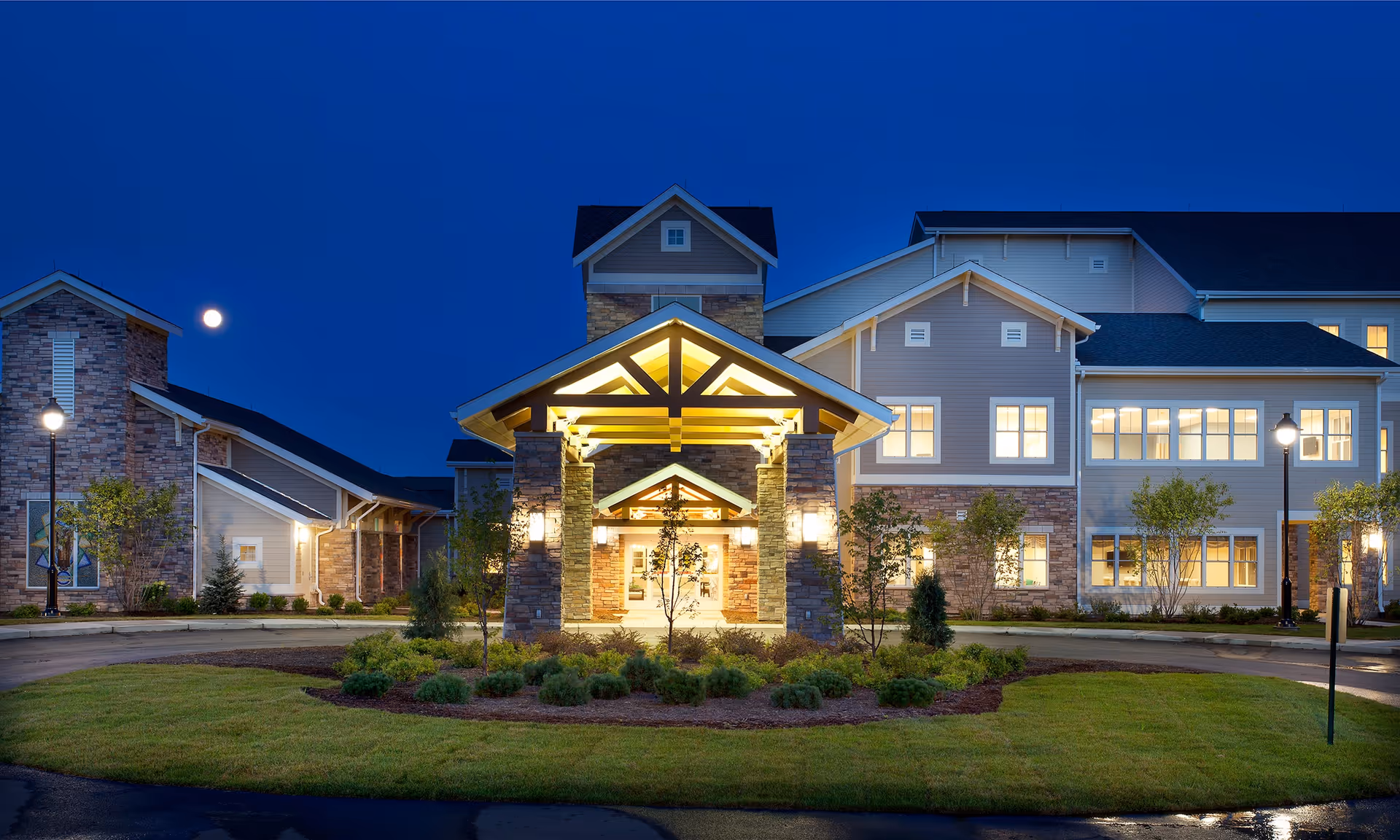 Exterior view of a senior living facility named Kahl Home at dusk, showing a well-lit entrance with stone pillars and a peaked roof, surrounded by landscaped greenery and a circular driveway under a clear evening sky with a visible moon.