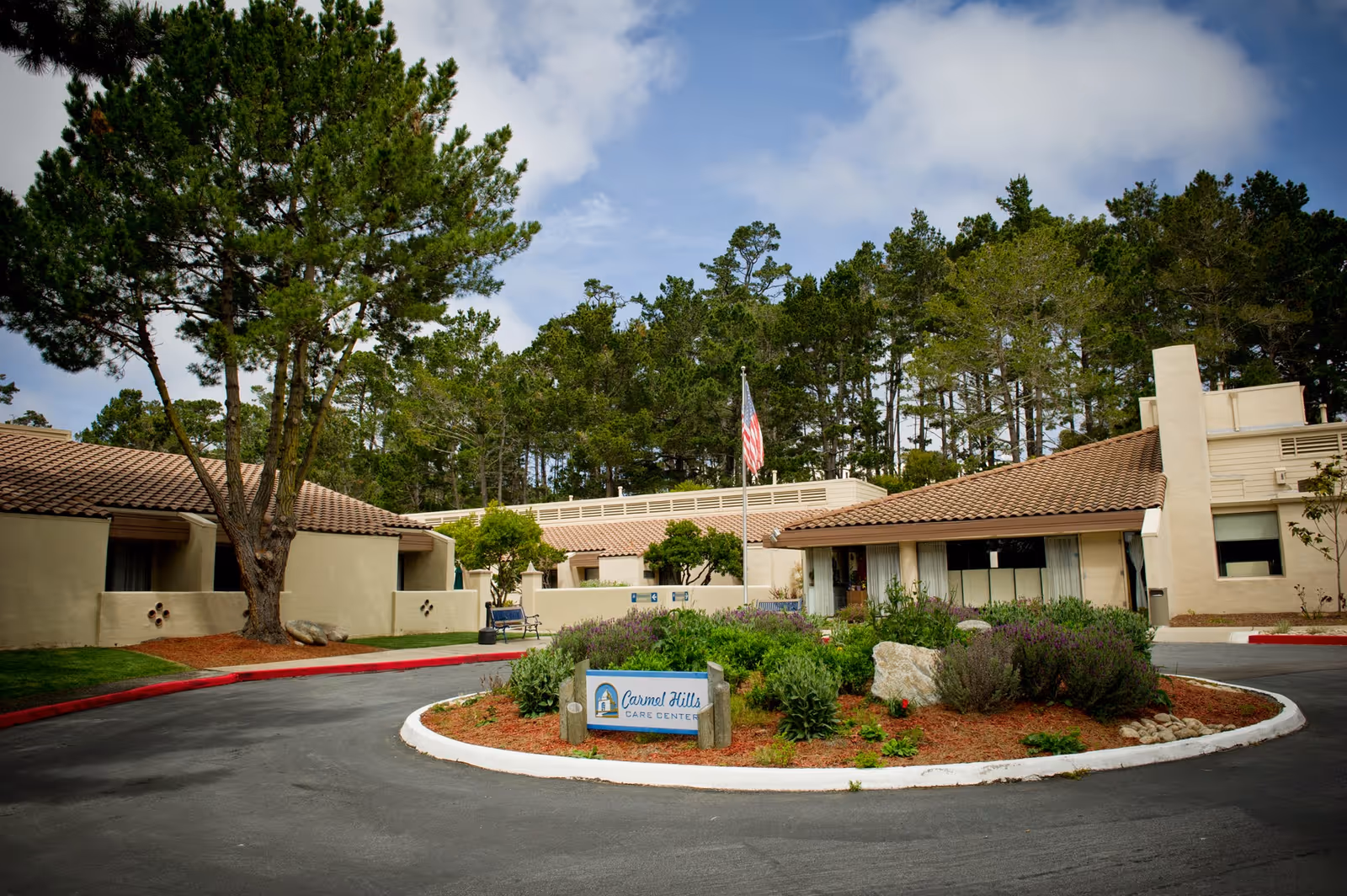 Exterior view of Carmel Hills Care Center showing a circular driveway with a landscaped island in the center, featuring plants and a sign with the facility's name. The building has beige walls and brown tiled roofs, surrounded by tall trees under a partly cloudy sky.