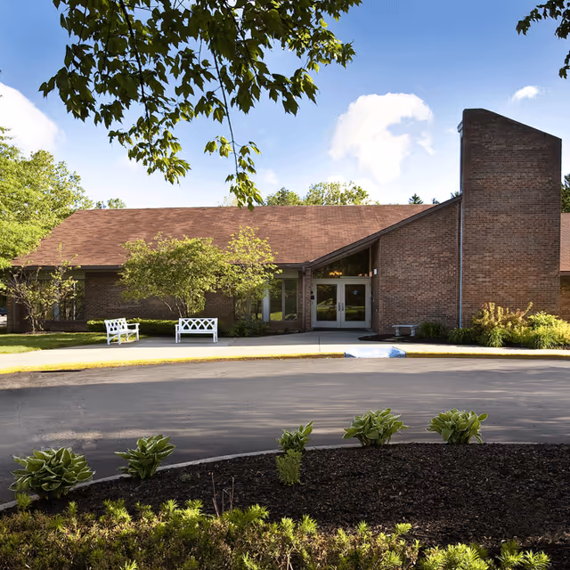Exterior view of a single-story brick building with a sloped roof, surrounded by greenery and small plants. There are two white benches near the entrance, which has double glass doors. The sky is partly cloudy with blue patches visible.
