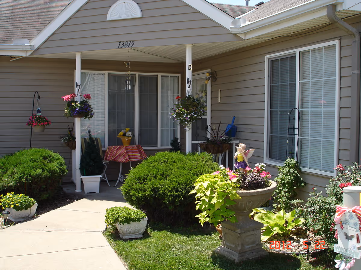 Front porch of a residential unit at Cottages of Kansas City East with beige siding, a small covered patio area with a table covered by a striped cloth, chairs, hanging flower baskets, potted plants, and well-maintained bushes and garden beds.