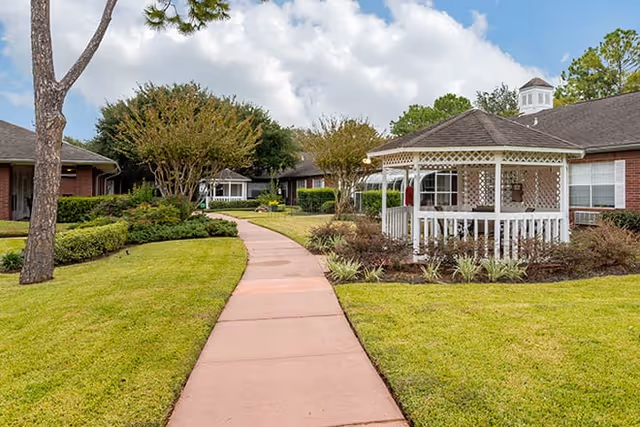 A paved walkway curves through a well-maintained garden area with green grass, shrubs, and trees. On the right side, there is a white gazebo with lattice panels, and in the background, there are single-story brick buildings with windows. The sky is partly cloudy.