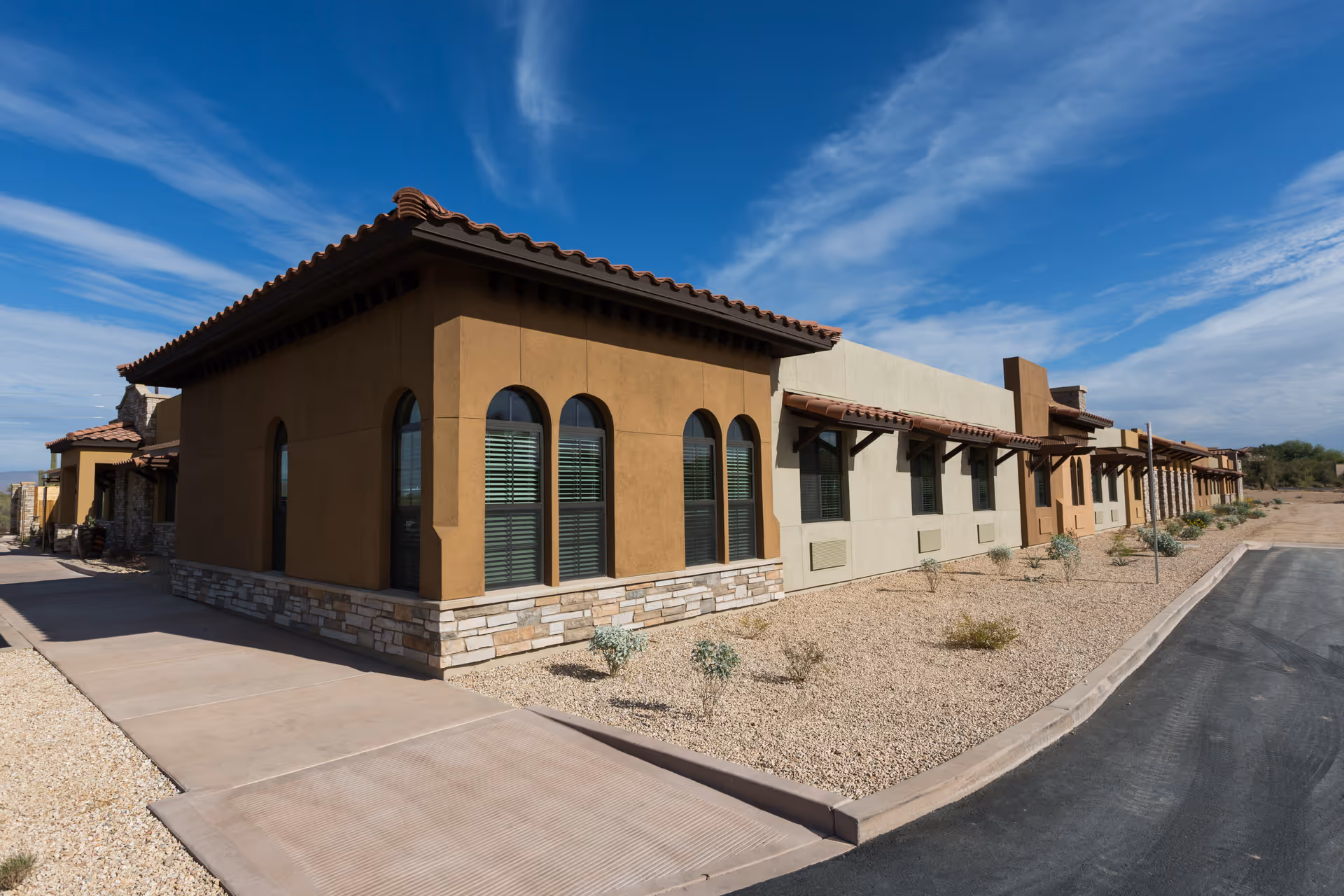 Exterior view of a single-story building with a southwestern architectural style, featuring arched windows, a tiled roof, and a stone base. The building is surrounded by a gravel landscape with small shrubs and a paved driveway under a blue sky with some clouds.