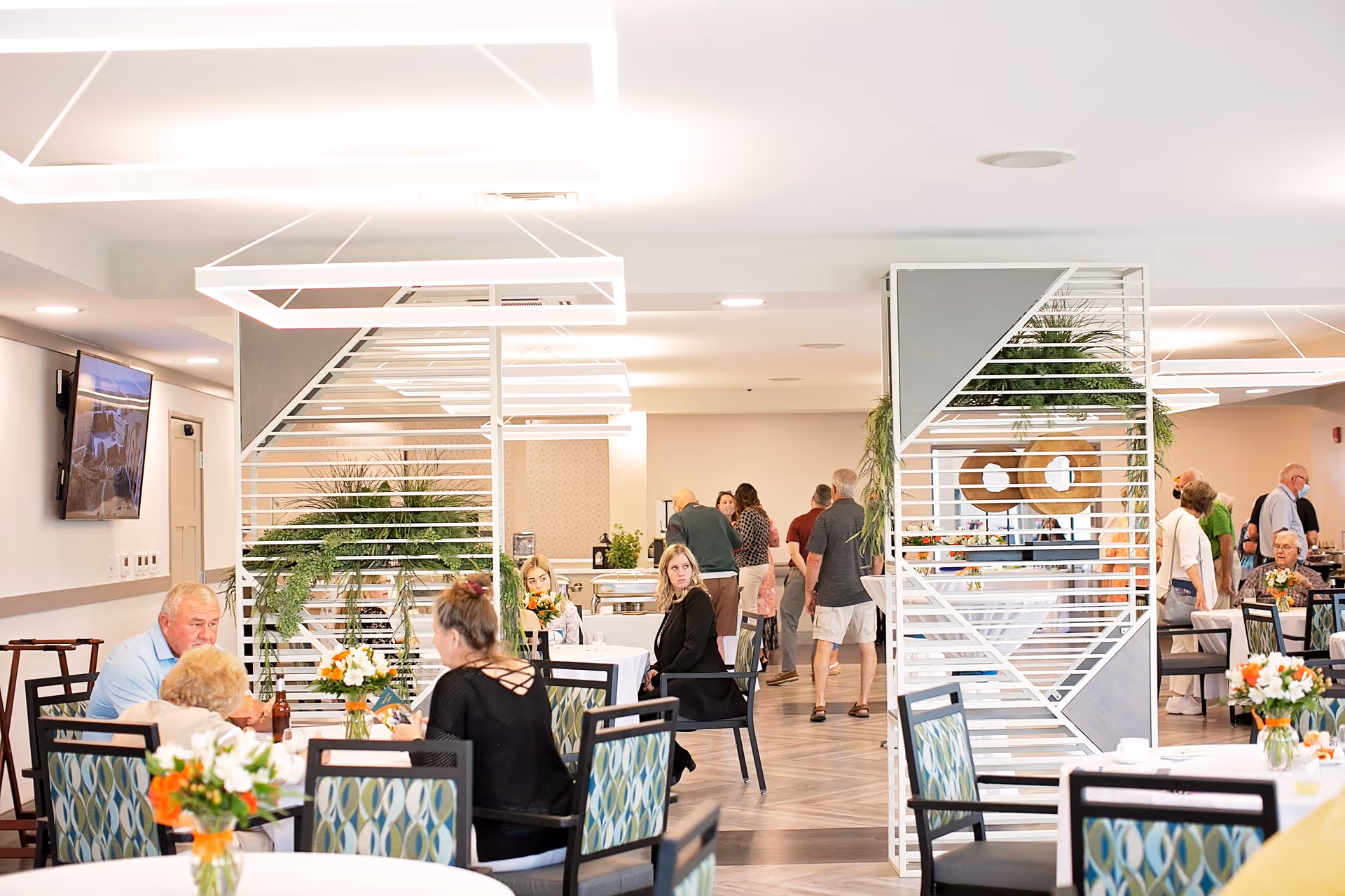 A bright dining area in a senior living facility with several tables and chairs. People are seated and engaged in conversation, while others are standing or walking in the background. The room features modern decor with white geometric light fixtures, decorative partitions with plants, and floral centerpieces on the tables.