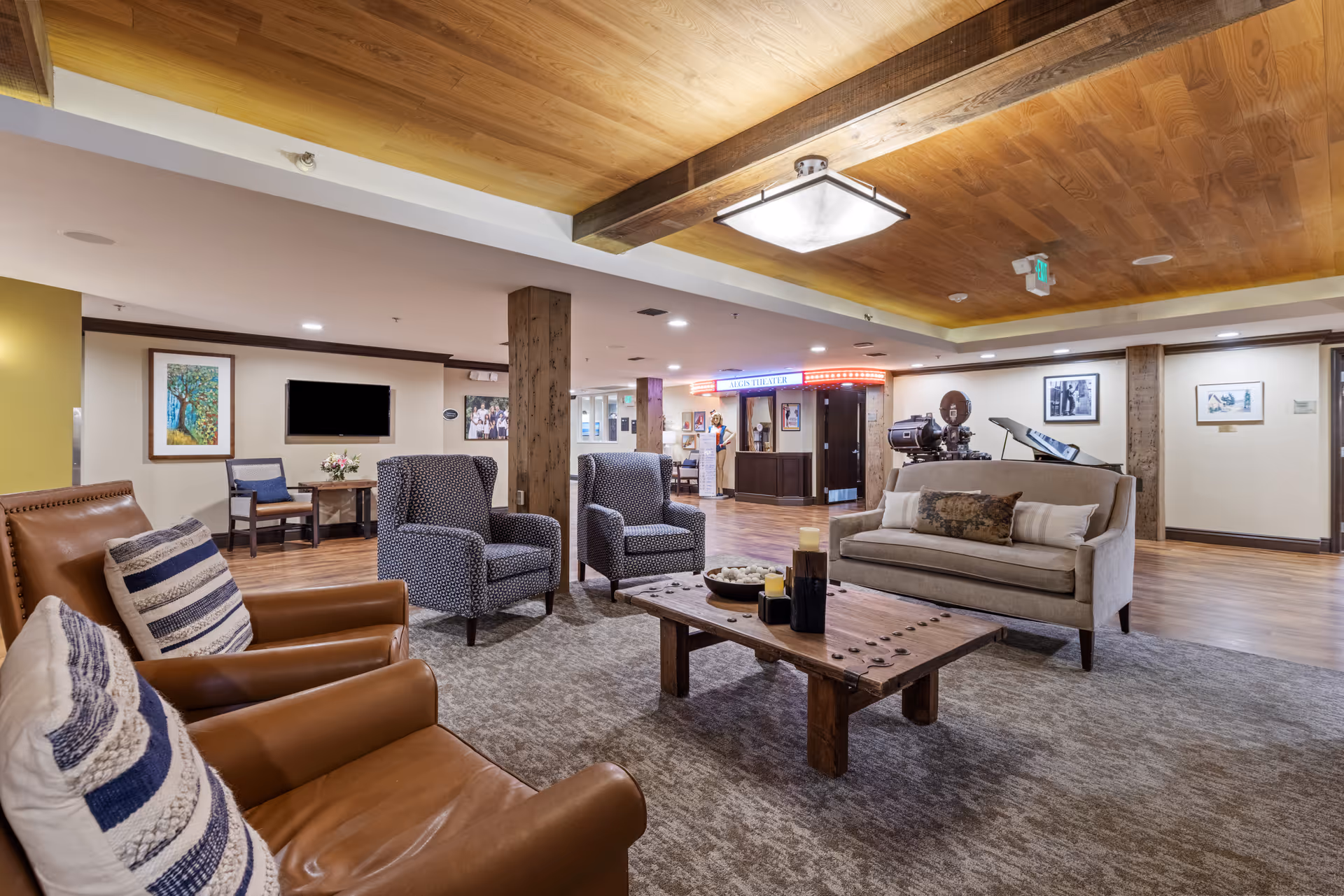 A spacious and well-lit living room area in a senior living facility with comfortable seating including two brown leather armchairs with striped pillows, two patterned armchairs, and a beige sofa with cushions. A wooden coffee table with decorative items sits on a large carpet. The room features wooden flooring, a wooden ceiling with beams, framed artwork on the walls, and a reception or service desk area in the background with a neon sign.