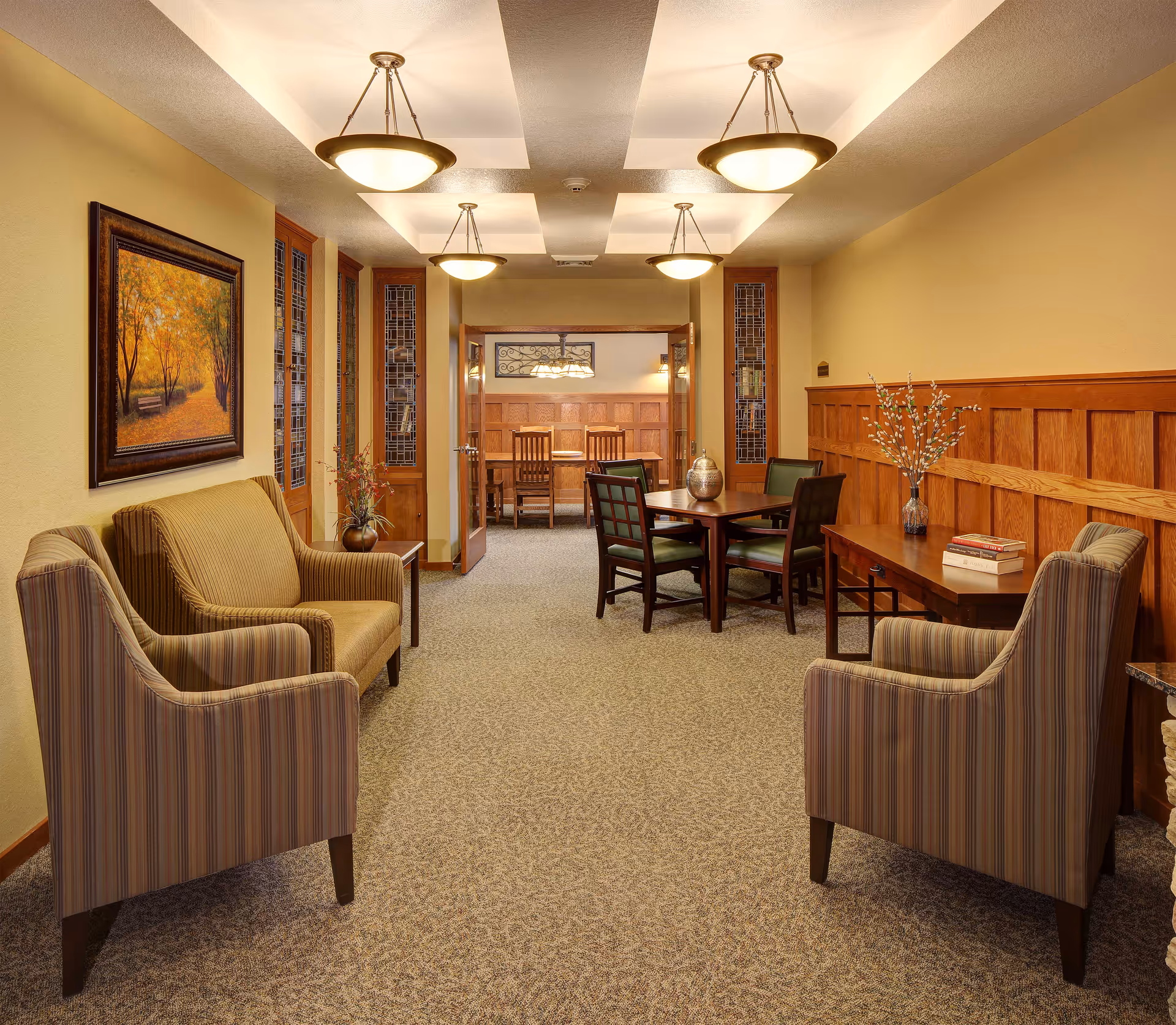 A cozy interior common area in Heritage Assisted Living & Memory Care featuring a seating arrangement with a striped armchair and a loveseat on the left, another striped armchair on the right, and a wooden table with four chairs in the center. The walls are painted beige with wooden paneling on the right side. There is a framed painting of a tree-lined path on the left wall and decorative glass panels flanking a set of double doors leading to another room with a wooden dining table and chairs. The ceiling has recessed lighting fixtures.