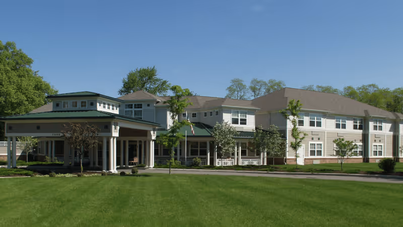Exterior view of a large, two-story senior living facility building with beige siding and a green roof. The building is surrounded by well-maintained green lawns, small trees, and shrubs under a clear blue sky.