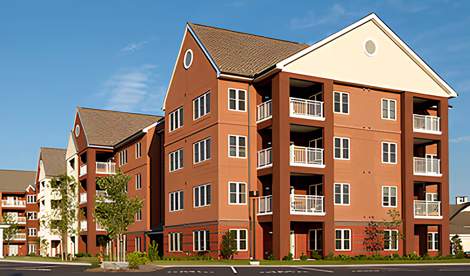 Exterior view of a multi-story senior living facility building with red and beige walls, multiple balconies, and several windows under a clear blue sky.