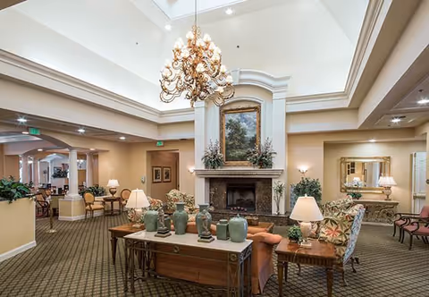 A spacious and elegantly decorated senior living facility common area with a high ceiling and large chandelier. The room features a central fireplace with a framed painting above it, surrounded by comfortable armchairs and sofas. Several tables with lamps and decorative vases are arranged around the seating area. The carpeted floor and warm lighting create a cozy atmosphere.