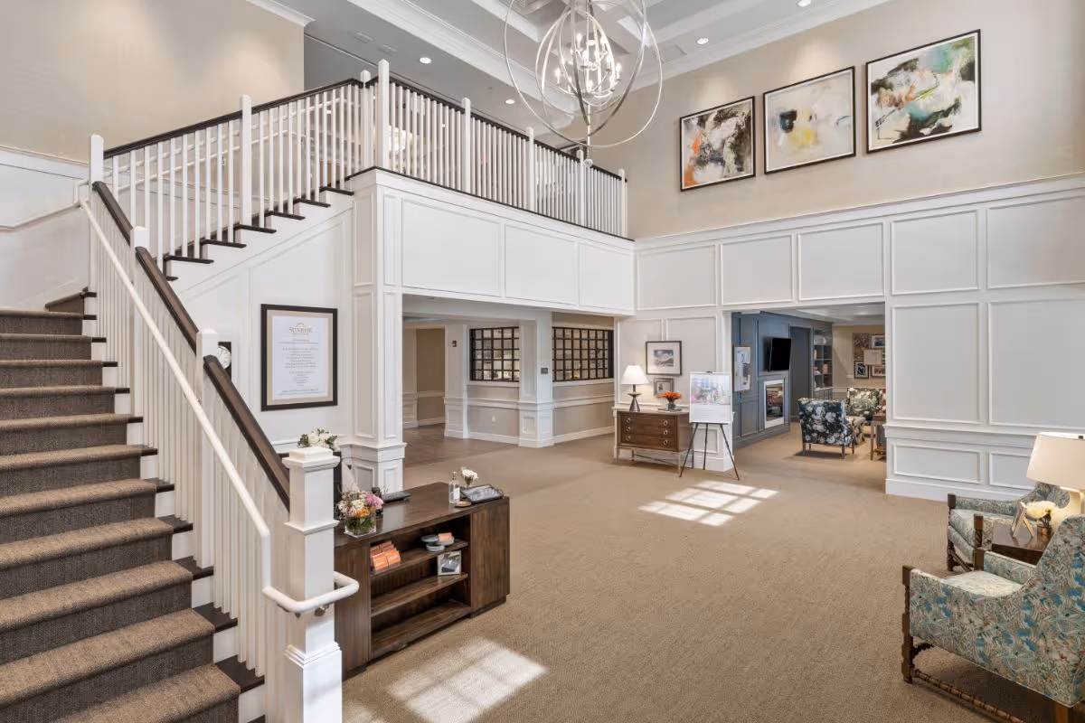 Spacious and well-lit interior common area of a senior living facility featuring a carpeted staircase with white railings, a wooden console table with flowers and books, a sitting area with patterned armchairs, a chandelier, and abstract artwork on the walls.