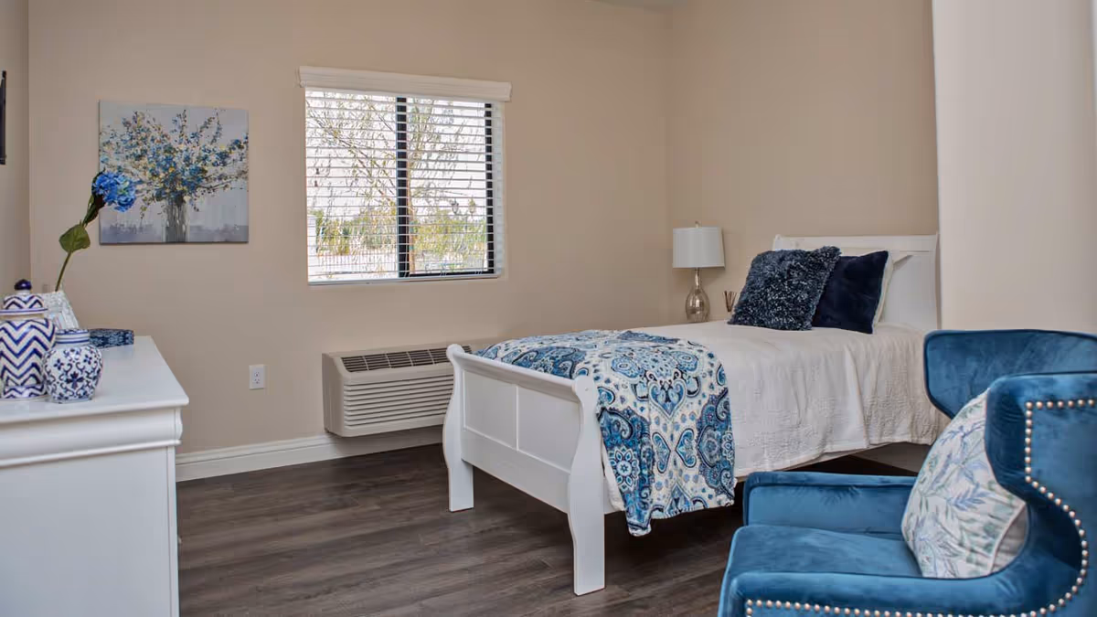 A neatly arranged bedroom with a white wooden bed featuring a blue and white patterned blanket and dark blue pillows. There is a blue upholstered chair with a decorative pillow in the foreground, a white dresser with blue and white decorative items on the left, a window with blinds, and a lamp on a bedside table. The walls are beige and the floor is dark wood.