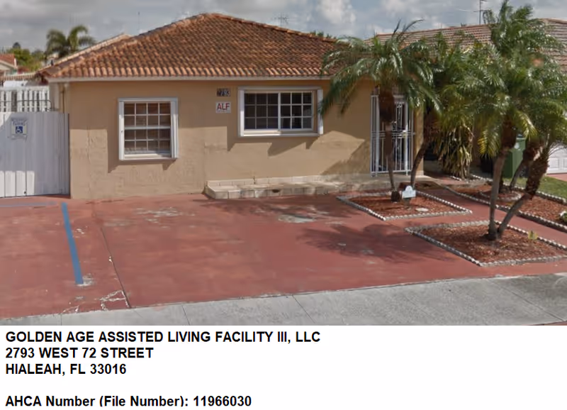 Front exterior view of a single-story assisted living facility building with beige walls and a red-tiled roof. There are two windows and a gated entrance. The driveway and parking area are paved in red, with a designated handicapped parking space marked by blue lines. Small landscaped areas with palm trees and decorative stones are visible near the entrance.