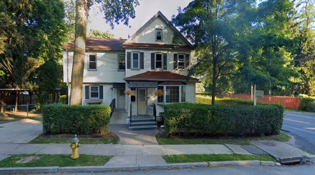 A two-story white house with a red roof and multiple windows, surrounded by green trees and bushes. There is a sidewalk and a yellow fire hydrant in front of the house, and a paved walkway leads to the front entrance with steps and a small porch.