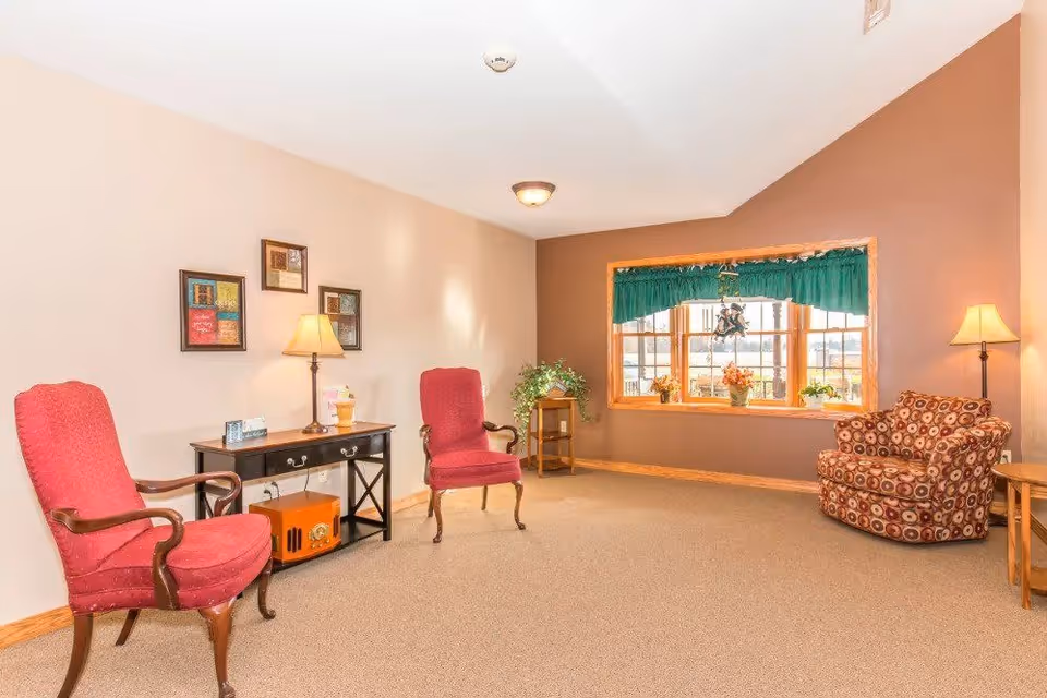 A cozy sitting area with two red upholstered armchairs, a patterned armchair, a wooden side table with a lamp, a small black console table with a lamp and decorative items, and a window with green valance curtains and potted plants on the windowsill.
