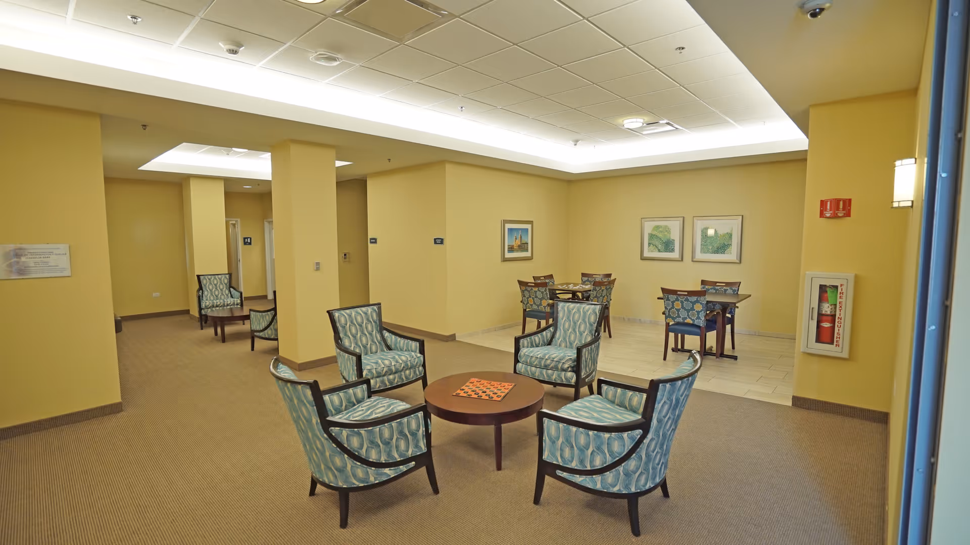 A common area in a senior living facility with four patterned armchairs arranged around a small round table with a checkerboard on it. In the background, there are two tables with chairs against the wall, which is decorated with framed artwork. The walls are painted yellow, and the floor is carpeted with a beige carpet, except for the area with the tables, which has a tiled floor. There is a fire extinguisher mounted on the wall to the right.