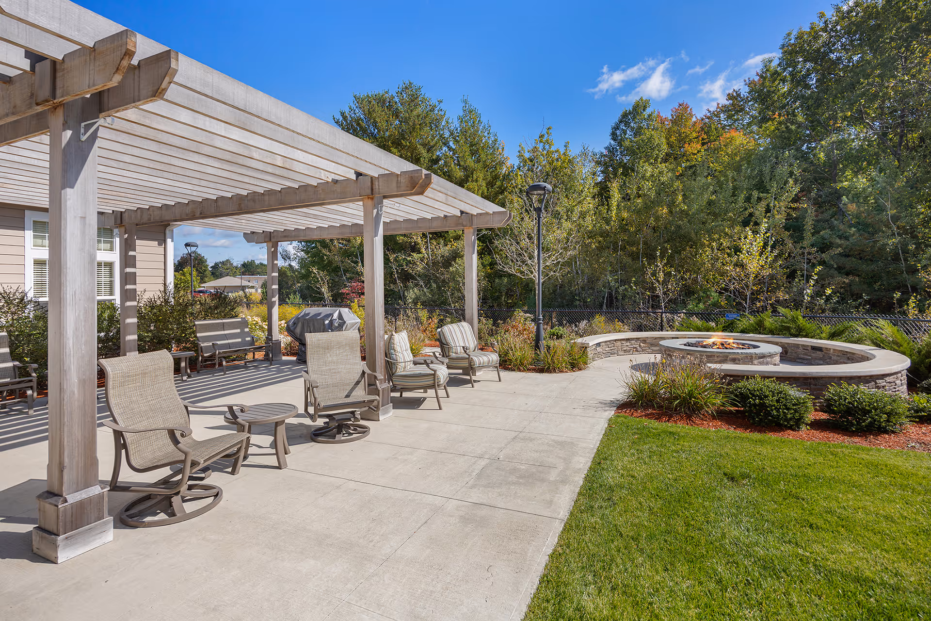 Outdoor patio area at Kennedy Meadows Gracious Retirement Living featuring a wooden pergola with several cushioned chairs and small tables underneath. There is a circular stone fire pit surrounded by a curved stone bench, with green grass and landscaped bushes nearby. Trees and a clear blue sky are visible in the background.