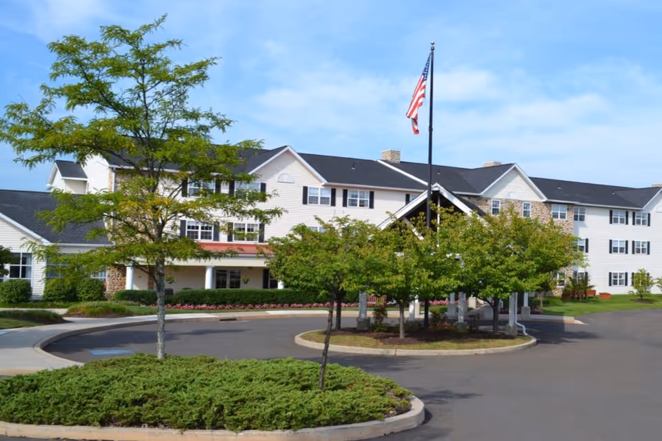 Exterior view of a large senior living facility building with white siding and black shutters. There is a circular driveway with landscaped greenery and trees in the center, and an American flag flying on a flagpole. The sky is clear and blue.