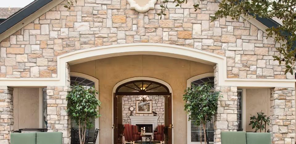 Exterior view of a stone building entrance with an arched doorway leading to a cozy interior featuring a fireplace, framed artwork, and red armchairs. Two potted trees flank the entrance, and green cushioned outdoor seating is visible in the foreground.