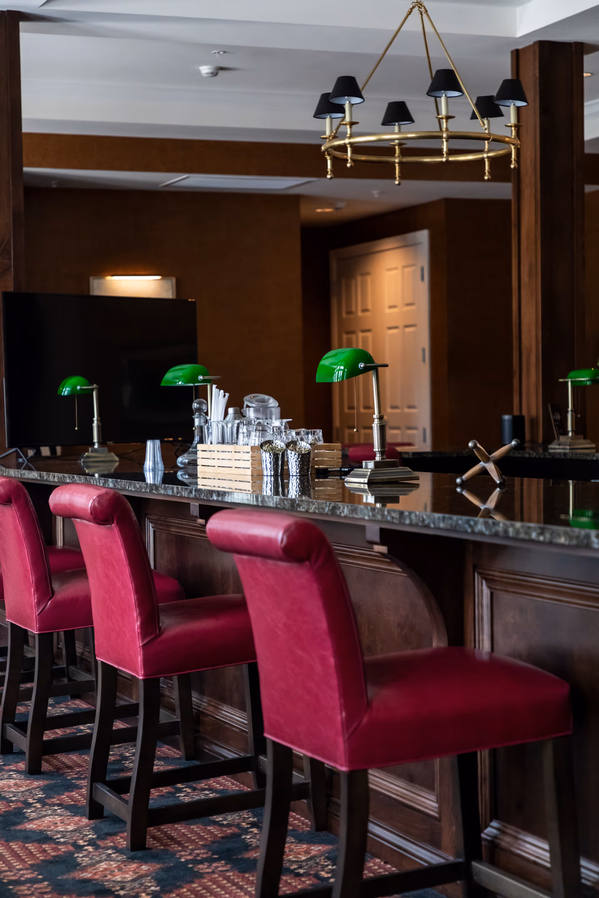 A bar area with a dark granite countertop and three red upholstered bar stools. On the counter are green banker-style lamps, a wooden crate holding glassware and utensils, and decorative items. The background features dark wood paneling, a flat-screen TV, and a white door. A brass chandelier with black lampshades hangs from the ceiling.