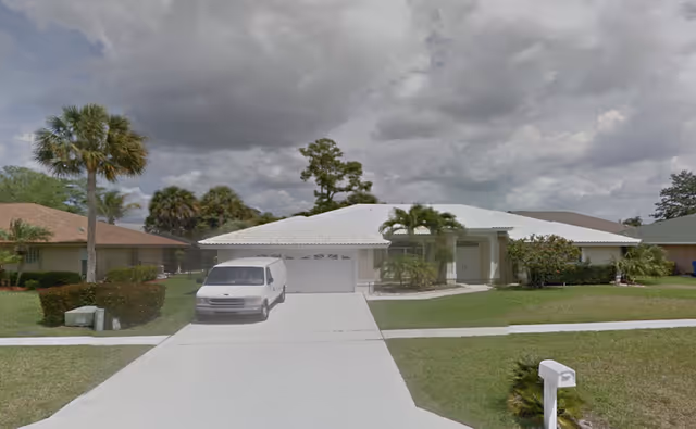 Single-story residential house with a white roof and garage, a white van parked in the driveway, palm trees and bushes in the front yard, and a cloudy sky overhead.