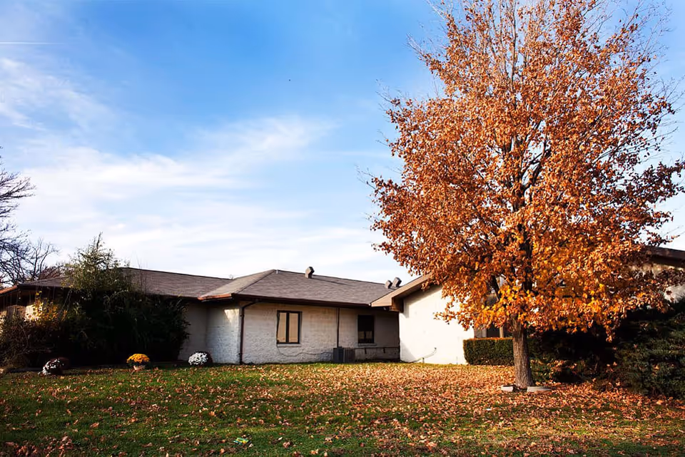 Exterior view of a single-story building with a brown roof and white walls, surrounded by a lawn with fallen autumn leaves and a large tree with orange leaves under a partly cloudy blue sky.