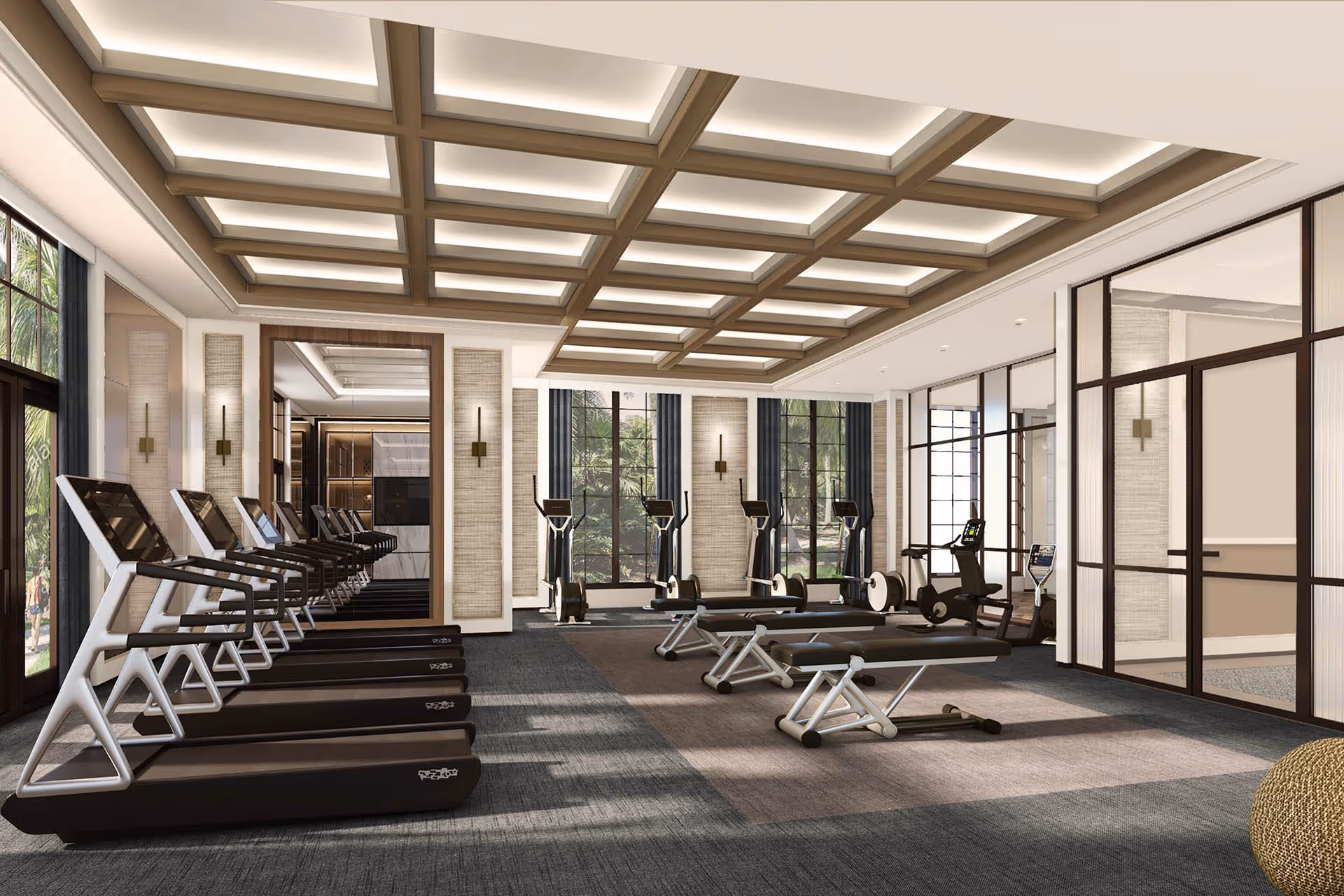 A modern fitness room with multiple treadmills lined up on the left side, exercise bikes and rowing machines near large windows, and two workout benches in the center. The room features a coffered ceiling with recessed lighting, large windows with views of greenery outside, and glass partitions on the right side.