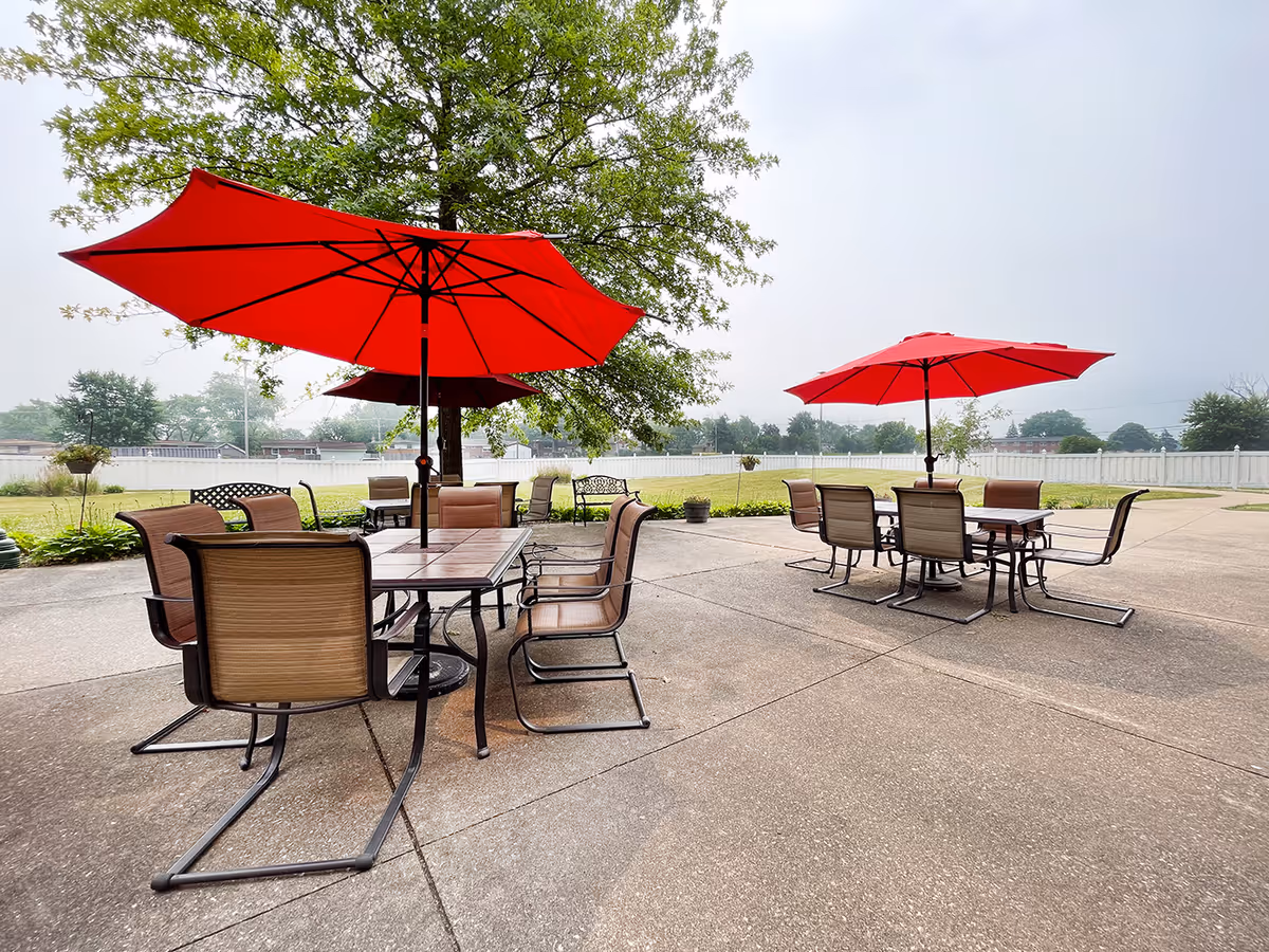 Outdoor patio area with two sets of tables and chairs, each shaded by a large red umbrella. The patio is surrounded by a white fence and greenery, with trees and a cloudy sky in the background.