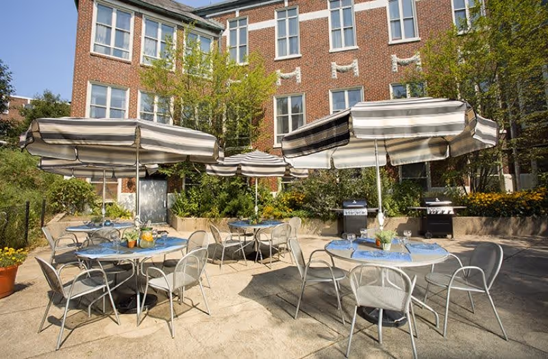 Outdoor patio area with round tables and metal chairs under large striped umbrellas. The tables are set with blue placemats, glasses, and a pitcher of lemonade. There are two grills in the background and a brick building with multiple windows behind the patio. Green trees and plants surround the area.
