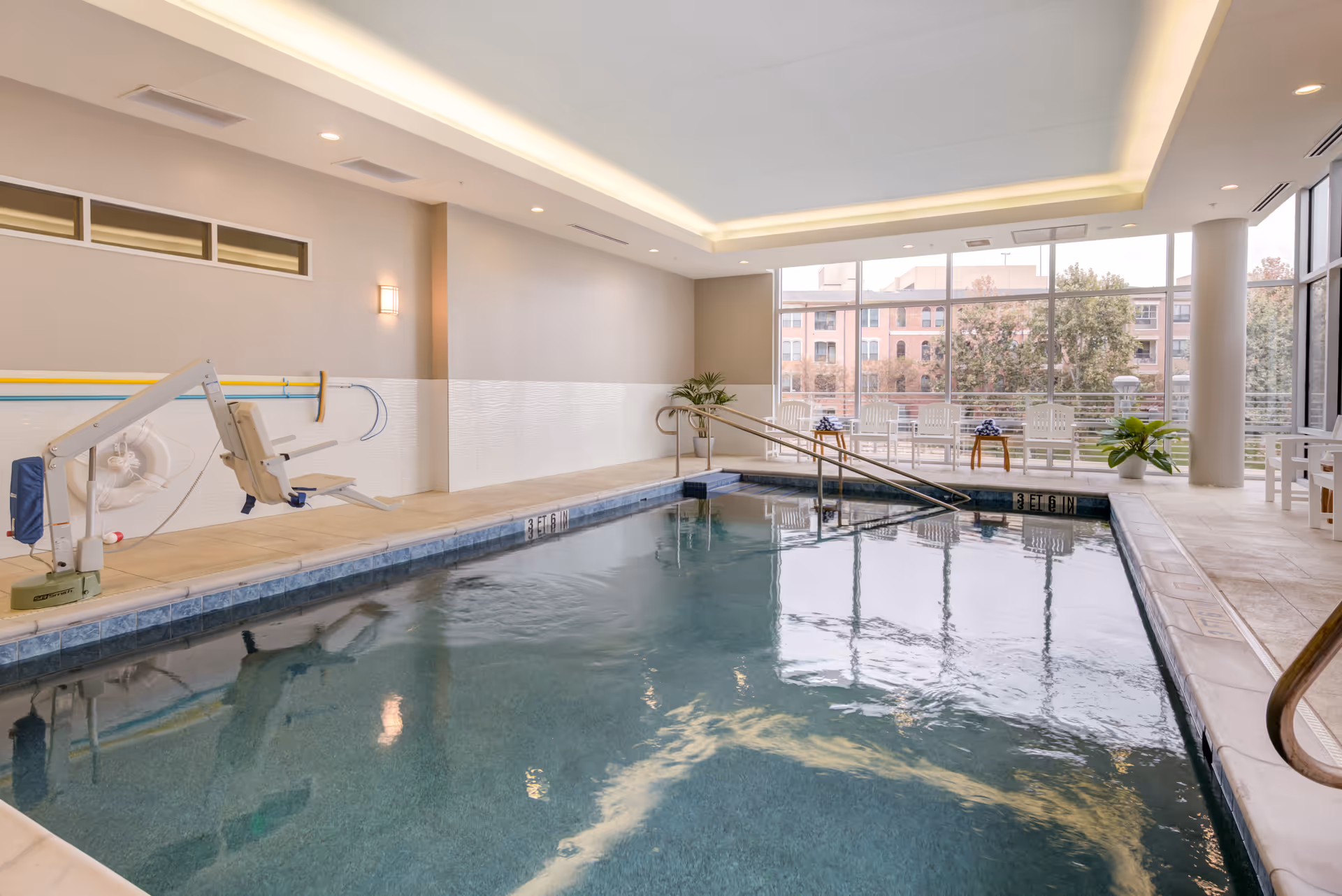 Indoor swimming pool with clear water, a pool lift for accessibility, handrails, and seating area with white chairs and small tables near large windows showing an outside view of buildings and trees.