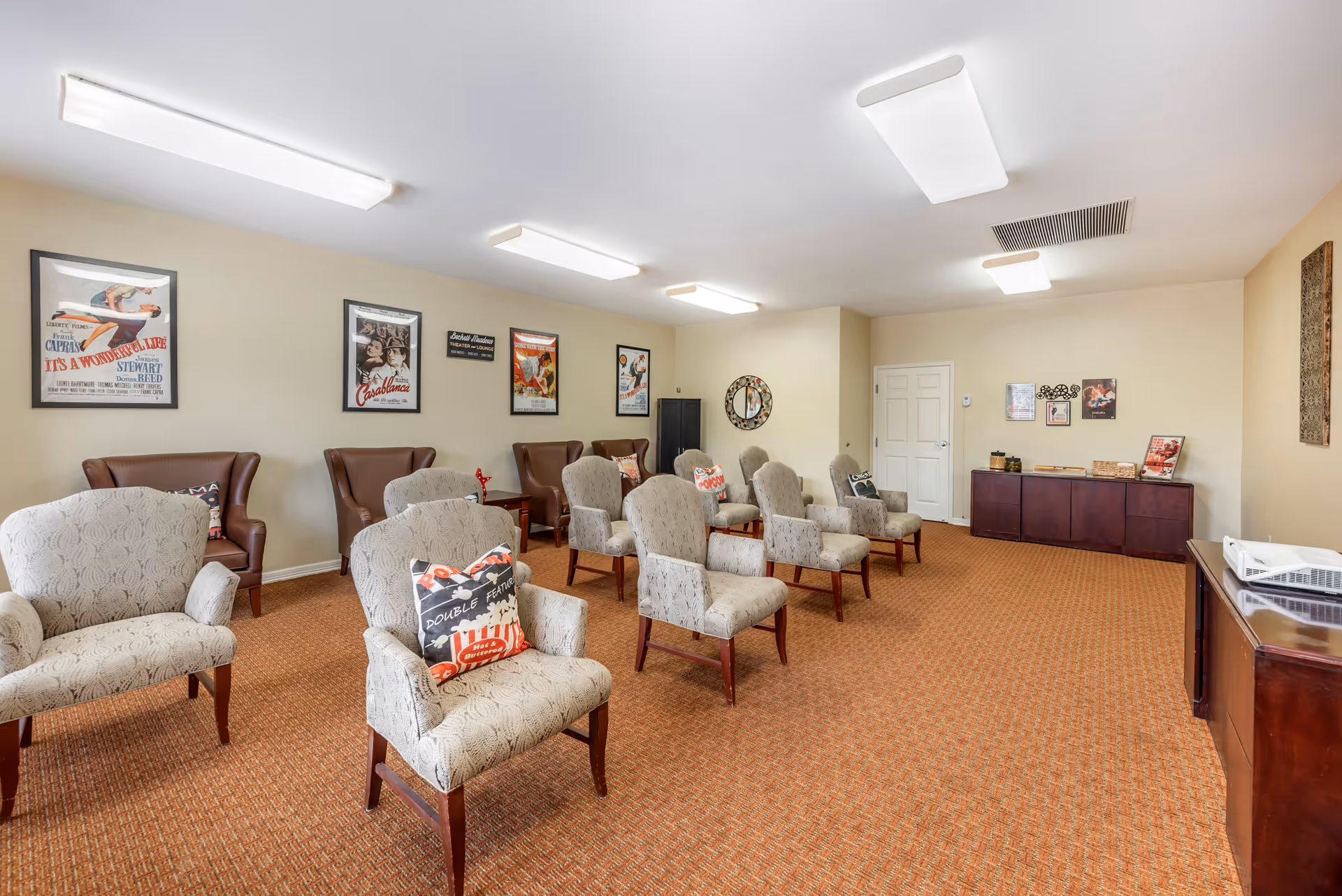 A small theater or lounge room with multiple upholstered chairs arranged in rows facing a projector on a wooden cabinet. The walls are decorated with vintage movie posters and a round decorative mirror. The room has beige walls, a patterned carpet, and fluorescent ceiling lights.