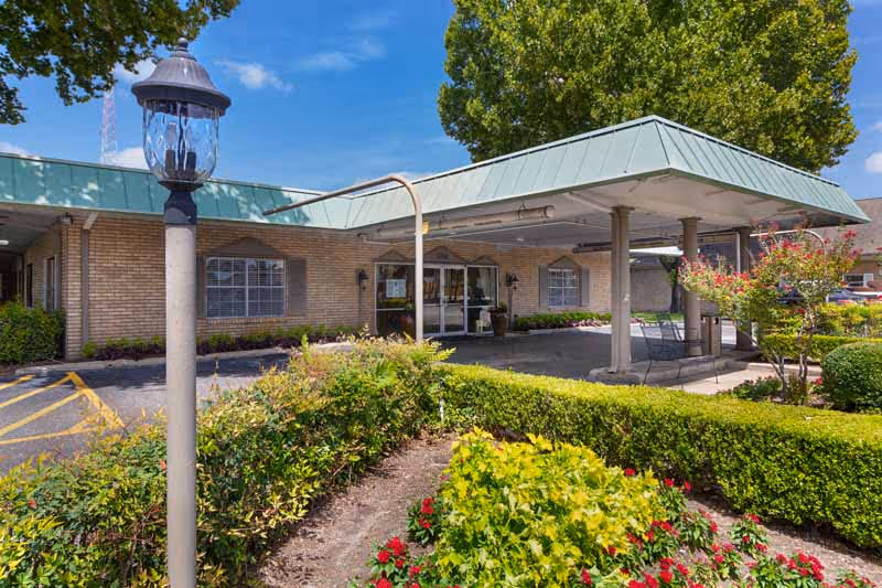 Exterior view of Memorial City Nursing and Rehabilitation Center showing the entrance with a covered drop-off area, surrounded by well-maintained landscaping including bushes, flowers, and a lamp post under a clear blue sky.
