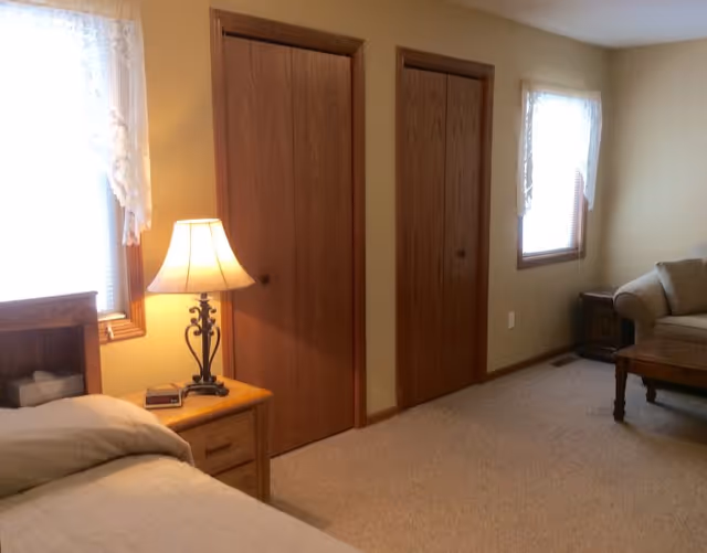 A cozy bedroom in a senior living facility with a bed partially visible on the left, a wooden nightstand with a lamp and clock, two closed wooden closet doors on the wall, two windows with lace curtains, a beige carpeted floor, and a beige couch with a wooden coffee table on the right side.