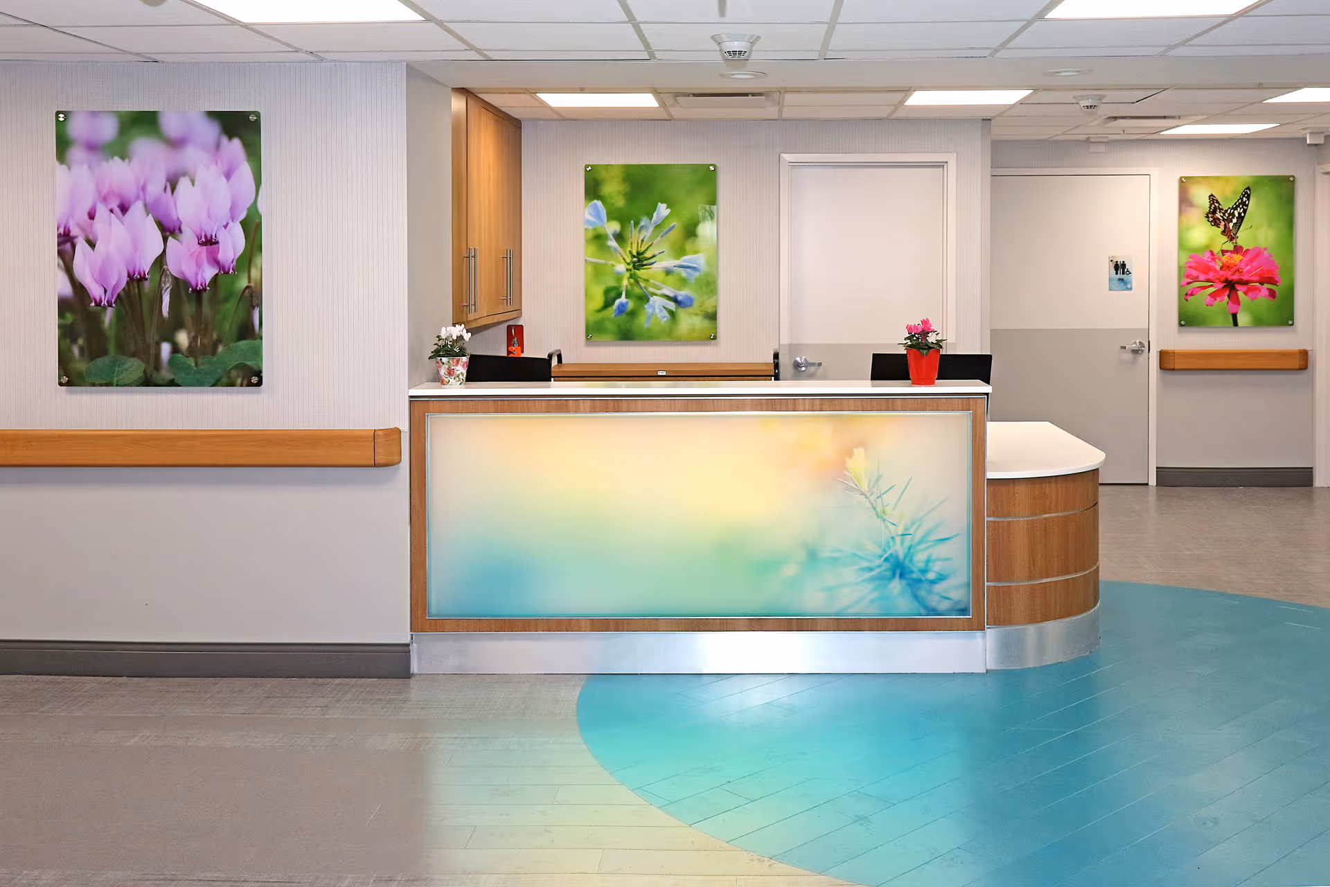 Reception desk area in a nursing and rehabilitation center with a wooden counter featuring a soft pastel floral design. Behind the desk are cabinets and two black chairs. The walls are decorated with three colorful flower and butterfly photographs. The floor has a blue and gray pattern, and the ceiling has recessed lighting.