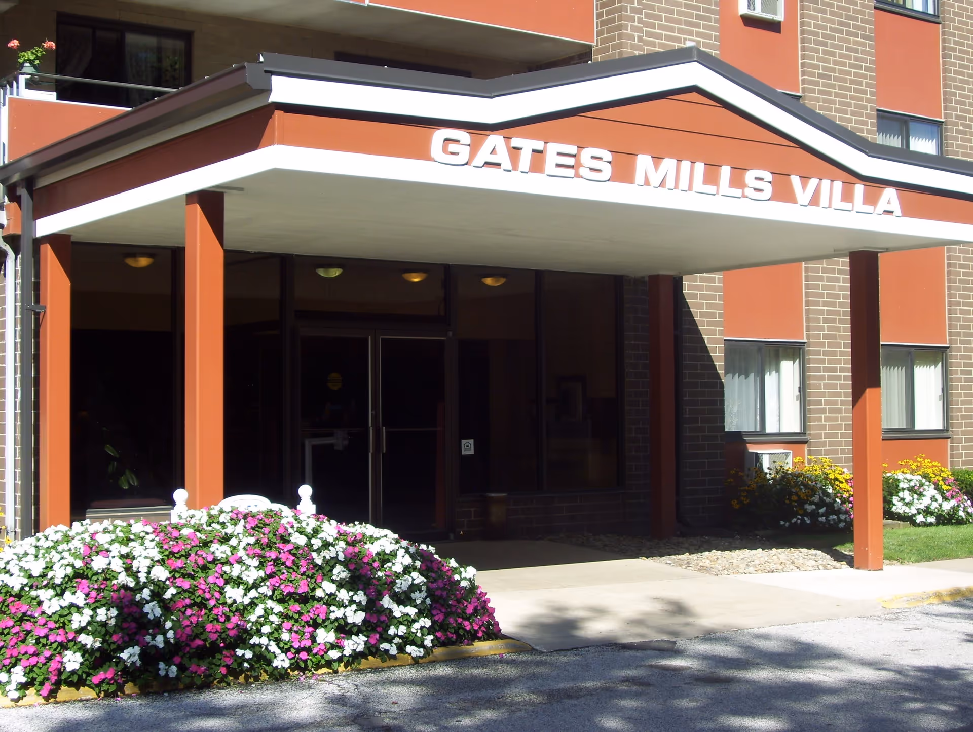 Entrance to Gates Mills Villa Apartments with a covered walkway supported by orange pillars. There are flower beds with white and purple flowers in front of the entrance and windows with curtains on the building facade.