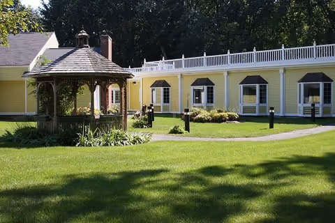 A well-maintained outdoor area of a senior living facility featuring a green lawn, a wooden gazebo with a shingled roof, and a yellow building with multiple windows and white railings in the background.
