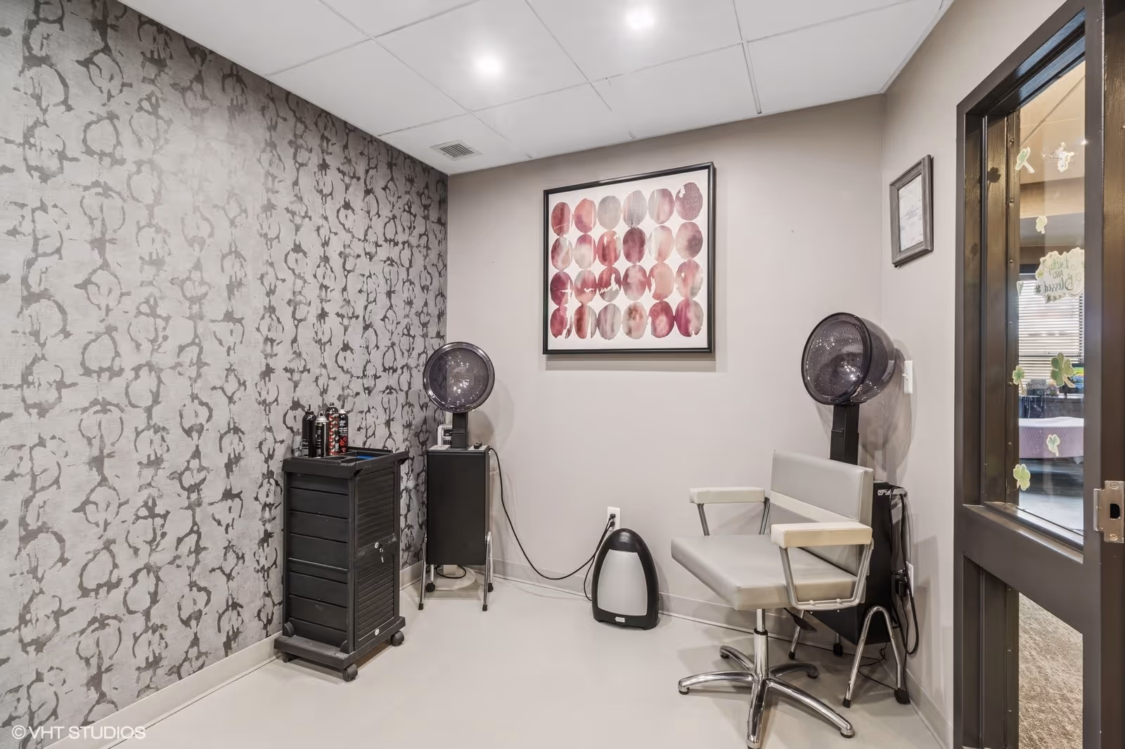 A small hair salon area with two hair drying chairs and two hair dryers mounted on stands. There is a black rolling cart with hair products on top, a patterned gray wallpaper on one wall, and a framed abstract artwork with circular pink and purple shapes on the adjacent wall. The floor is light-colored, and a door with glass panels is partially visible on the right side.