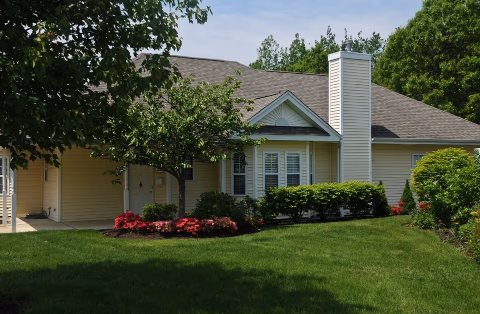 Exterior view of a single-story beige building with a gray shingled roof, surrounded by green grass, bushes, and trees under a partly cloudy sky.