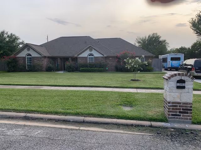 Single-story brick house with a gray shingled roof, surrounded by a well-maintained lawn and small trees. A stone and brick mailbox with the number 12802 is visible in the foreground on the right side. Several vehicles, including a blue bus and a black SUV, are parked near the house. The sky is partly cloudy with soft lighting.