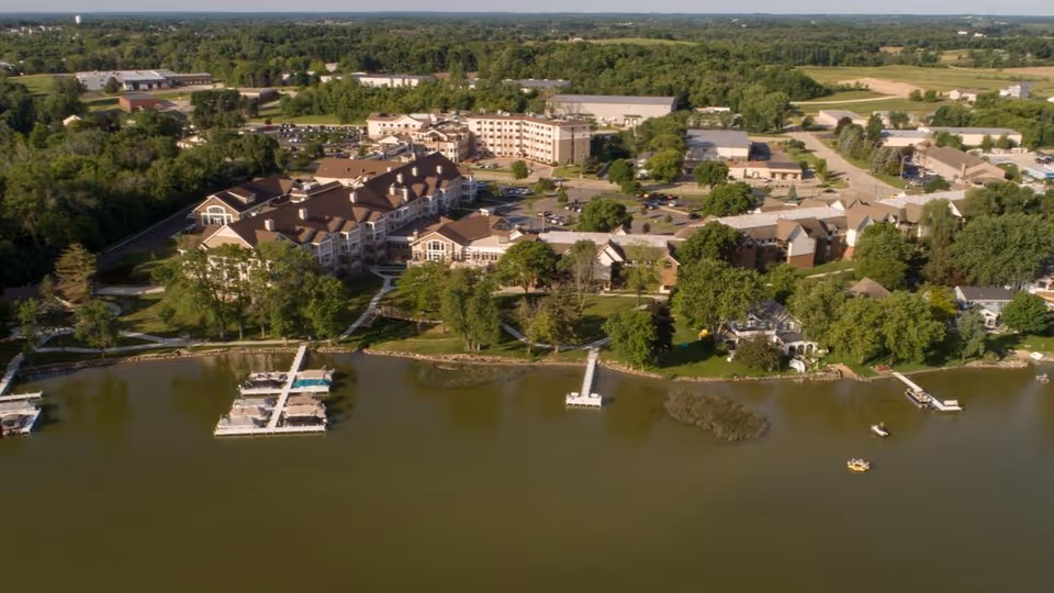 Aerial view of a senior living facility named Shorehaven situated along a waterfront with docks and boats. The facility consists of multiple buildings surrounded by trees and greenery, with parking areas and roads visible in the background.