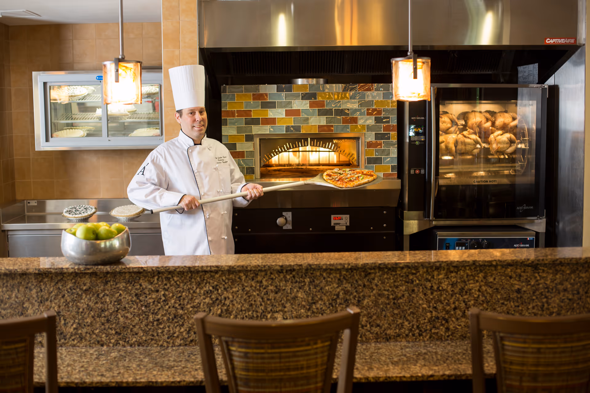 A chef in a white uniform and tall white hat is holding a pizza peel with a freshly baked pizza in a kitchen. Behind him is a wood-fired pizza oven with colorful tiles and a rotisserie oven with chickens roasting inside. A bowl of green apples is on the granite countertop in the foreground, with chairs lined up along the counter.