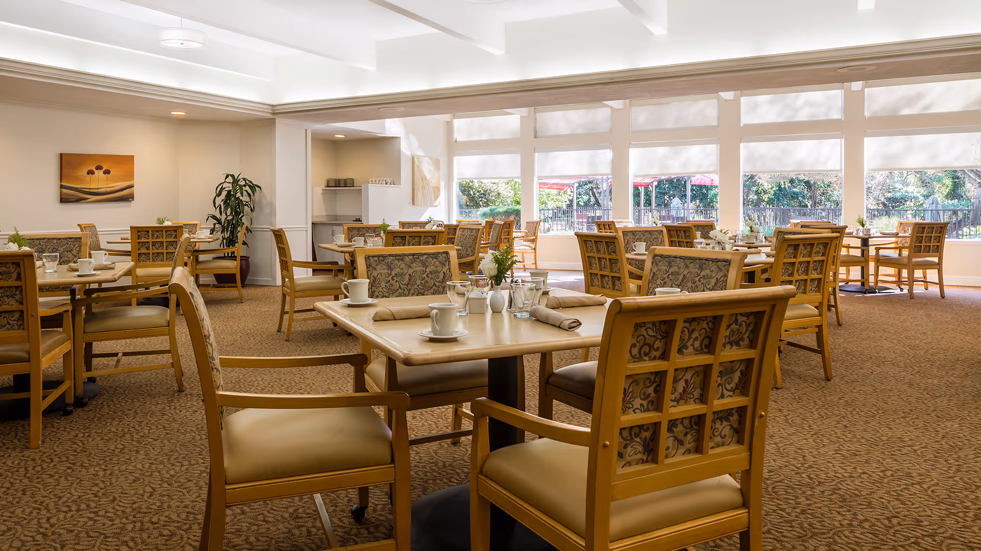 A bright and spacious dining room with multiple wooden tables and chairs arranged neatly. Each table is set with cups, glasses, napkins, and small flower vases. Large windows along one wall allow natural light to fill the room and offer a view of greenery outside. The room has a warm and inviting atmosphere with carpeted floors and light-colored walls.