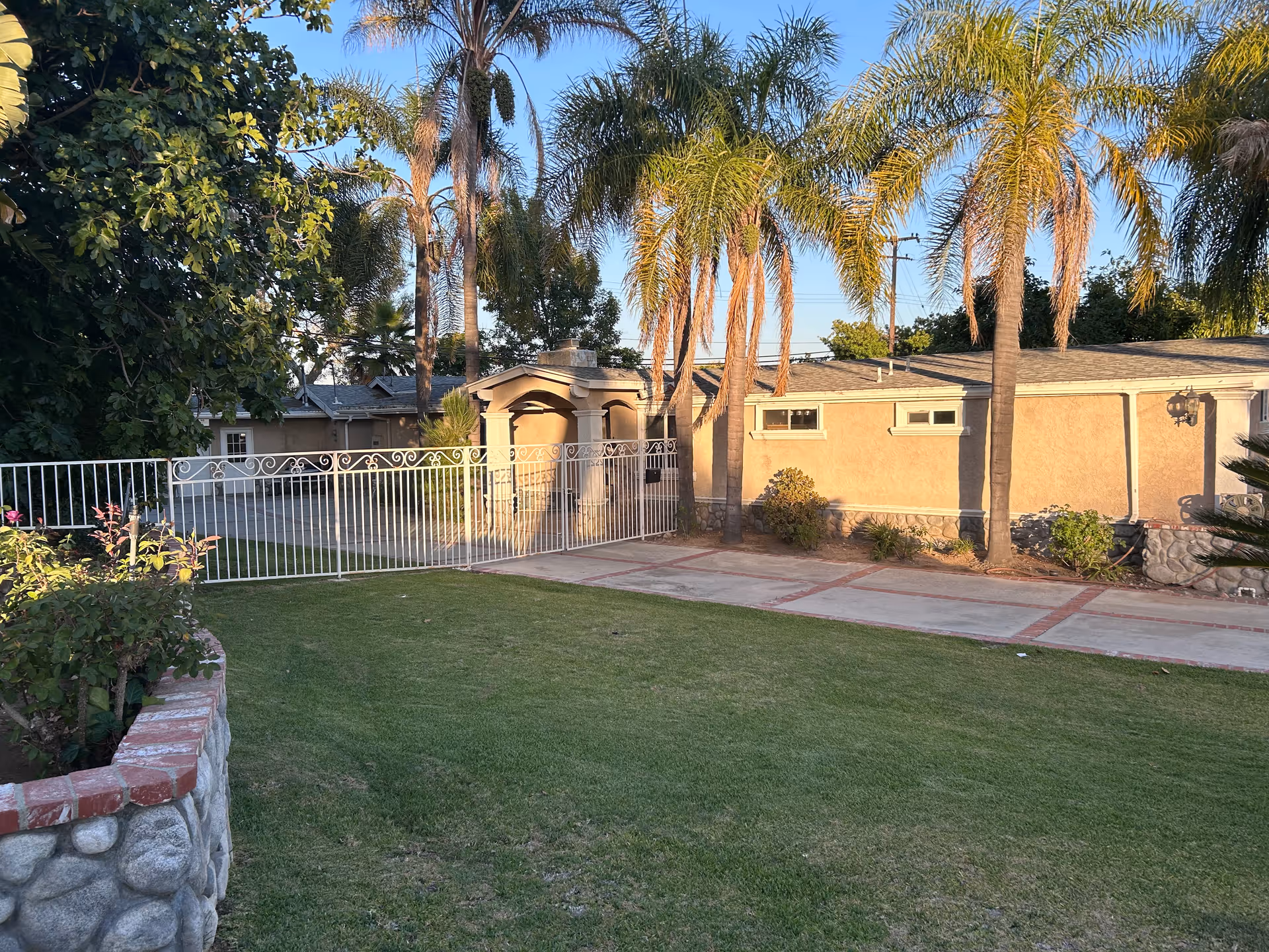 A sunny outdoor area with a green lawn, palm trees, and a beige building with small windows. There is a white metal fence and gate, along with a stone and brick planter on the left side.
