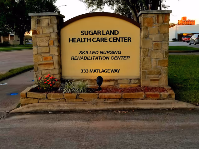 Stone sign at the entrance of Sugar Land Health Care Center, a skilled nursing rehabilitation center located at 333 Matlage Way, with some small plants and flowers at the base of the sign.