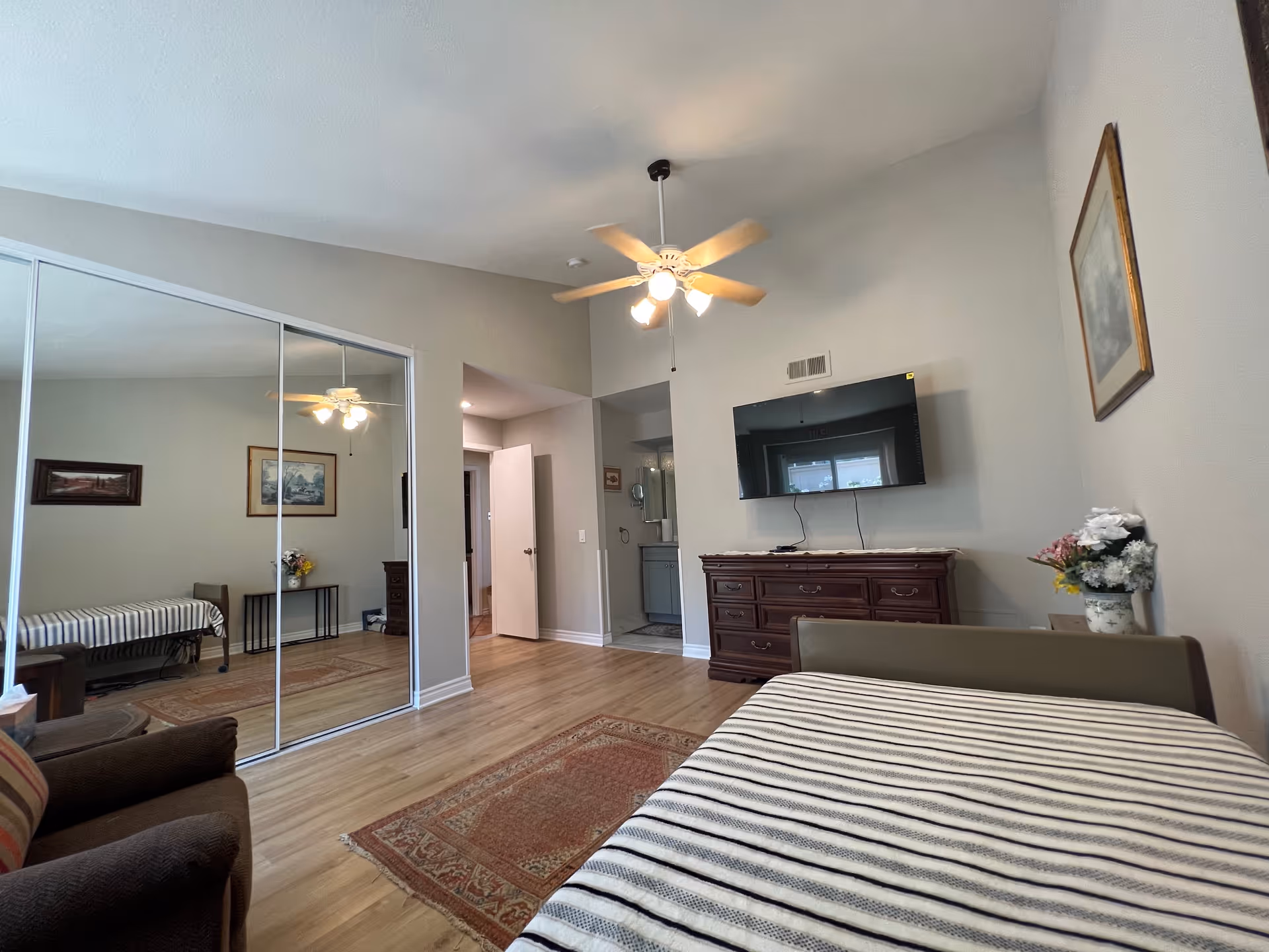 A bedroom in a senior living facility with a bed covered in a striped blanket, a brown armchair, a wooden dresser with a flat-screen TV mounted above it, a ceiling fan with lights, a large mirrored closet, and framed artwork on the walls. The room has light-colored walls and wood flooring with a small area rug.
