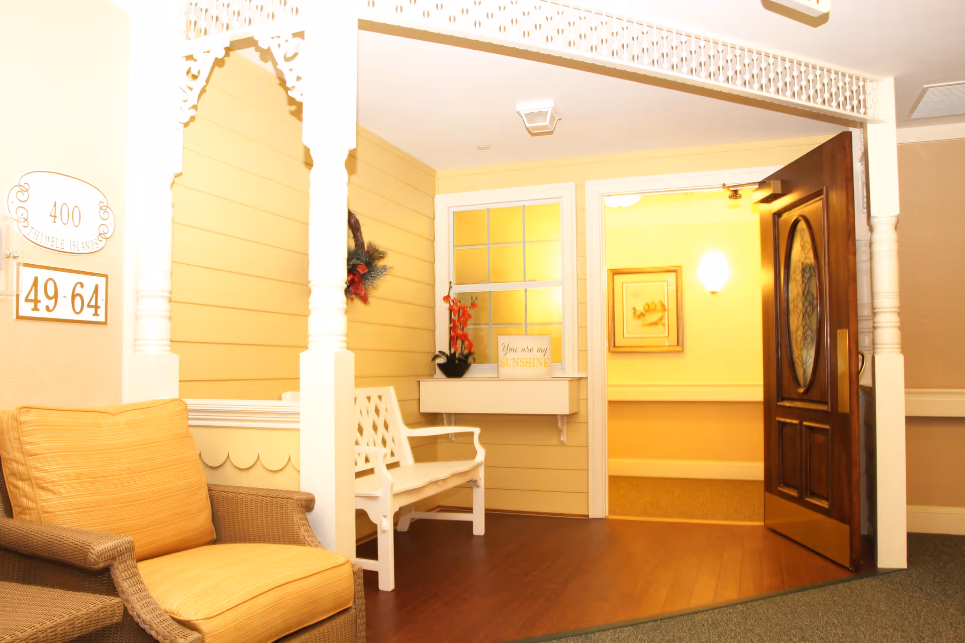 A cozy indoor seating area with a wicker armchair with a yellow cushion and a white wooden bench. The walls are painted yellow with white trim and decorative molding. A wooden door with an oval glass window is open, leading to a warmly lit hallway. There is a small shelf under a window with a potted plant and a sign that reads 'You are my SUNSHINE'. A sign on the wall indicates room numbers 49-64 and the number 400.