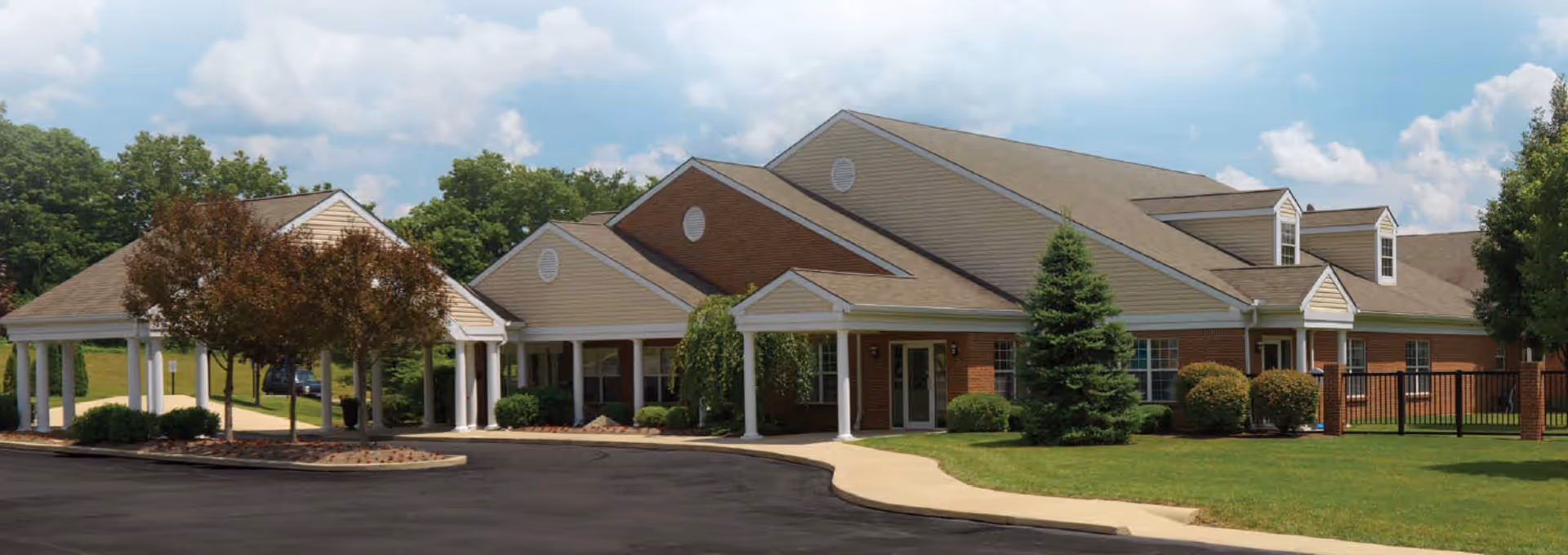 Exterior view of Hillspring Skilled Nursing Facility showing a single-story building with a covered entrance, surrounded by green lawns, trees, and a paved driveway under a partly cloudy sky.