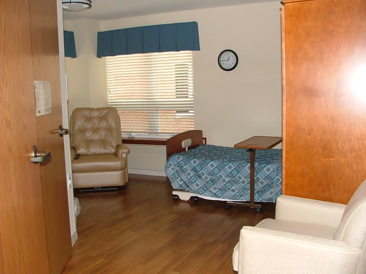 A small room with a hospital-style bed covered in a blue patterned blanket, a beige recliner chair near a window with closed blinds and a blue valance, a wooden cabinet, a beige armchair, and a wall clock showing the time as 2:10.