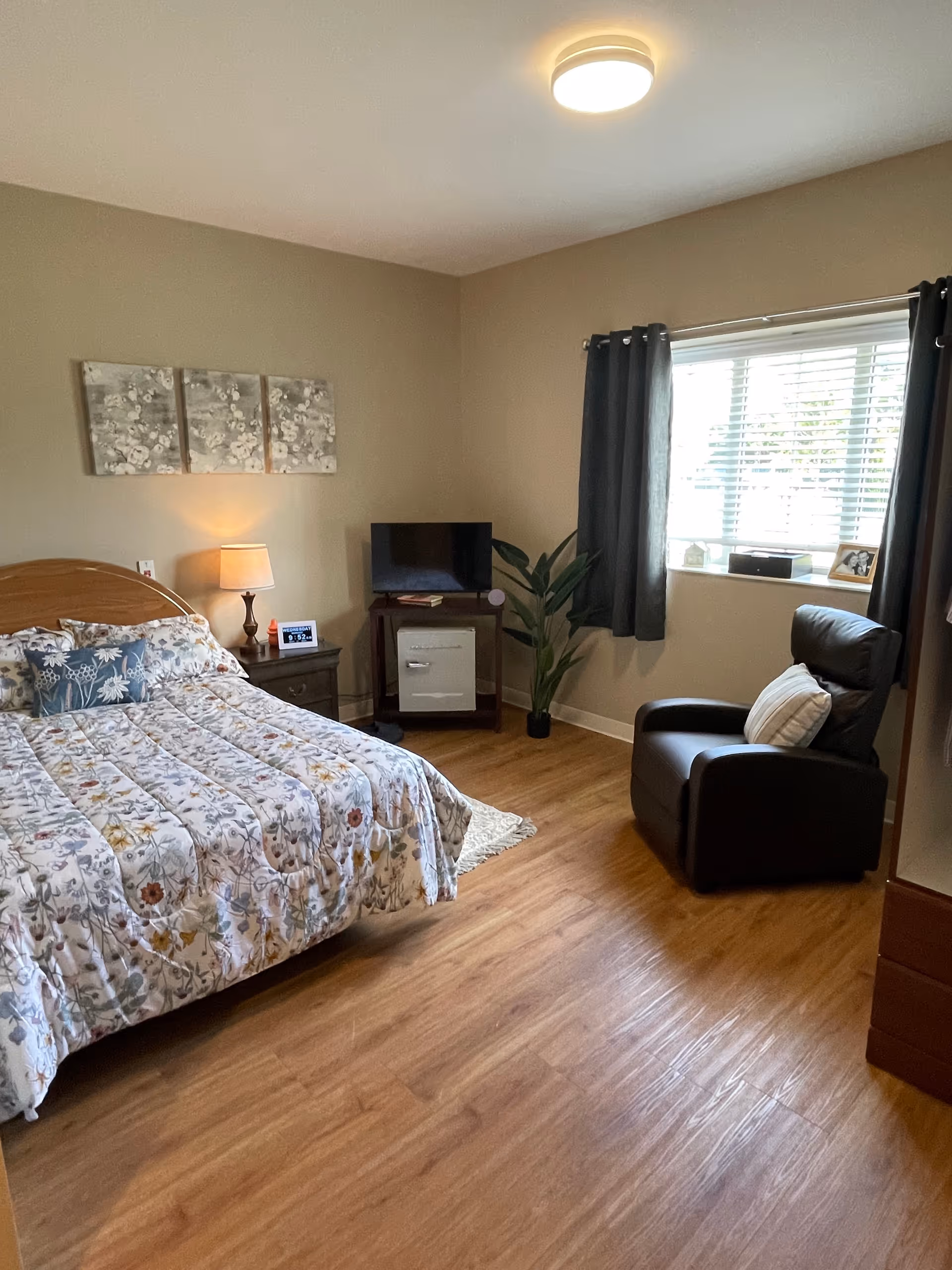 A cozy bedroom with a bed covered in a floral patterned comforter, a wooden headboard, a nightstand with a lamp and clock, a small TV on a stand with a plant beside it, a window with dark curtains, and a black armchair with a pillow.