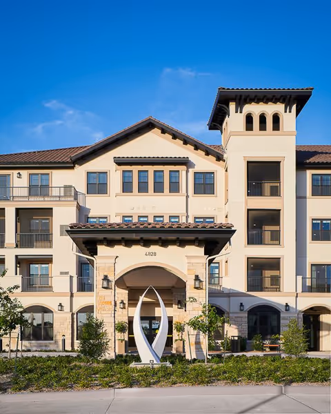 Front exterior view of a multi-story senior living facility with beige walls, a tiled roof, balconies, and a modern sculpture in front of the entrance. The building number 4828 is visible above the entrance archway.
