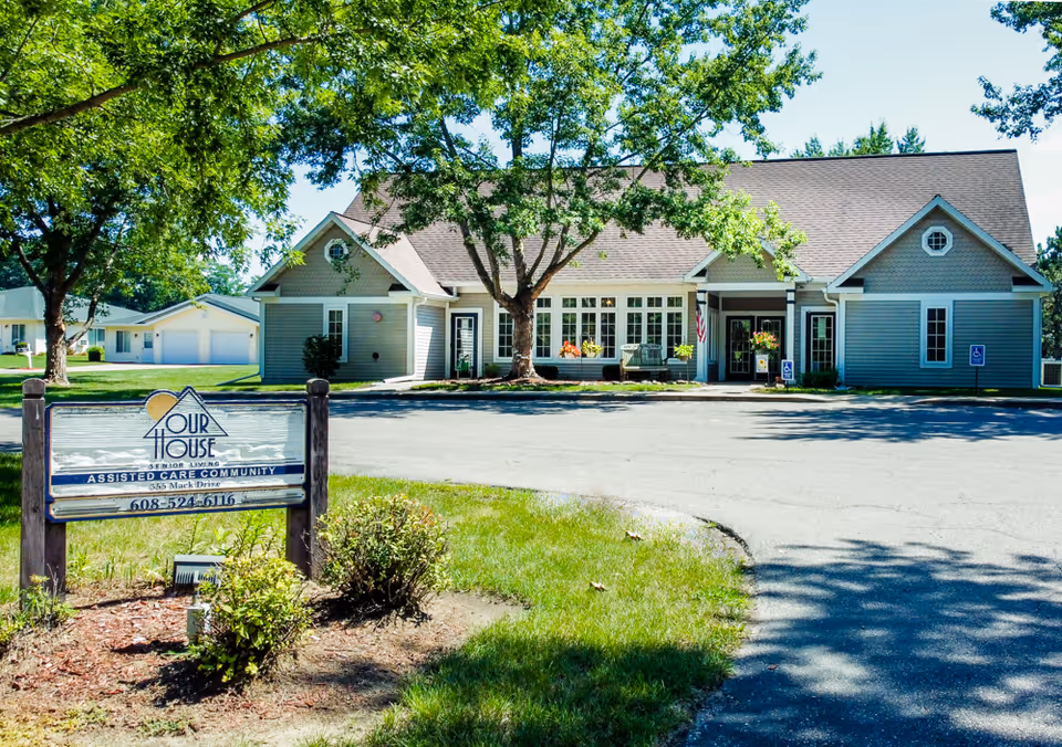 Exterior view of Our House Senior Living - Reedsburg Assisted Care facility showing a single-story building with a gable roof, surrounded by trees and greenery. There is a sign in the foreground with the facility's name and contact information. The area is well-maintained with a paved driveway and parking spaces.